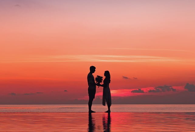 A romantic scene of a couple standing on a beach, looking into each other’s eyes, symbolizing the topic of manifesting someone to like you back. 